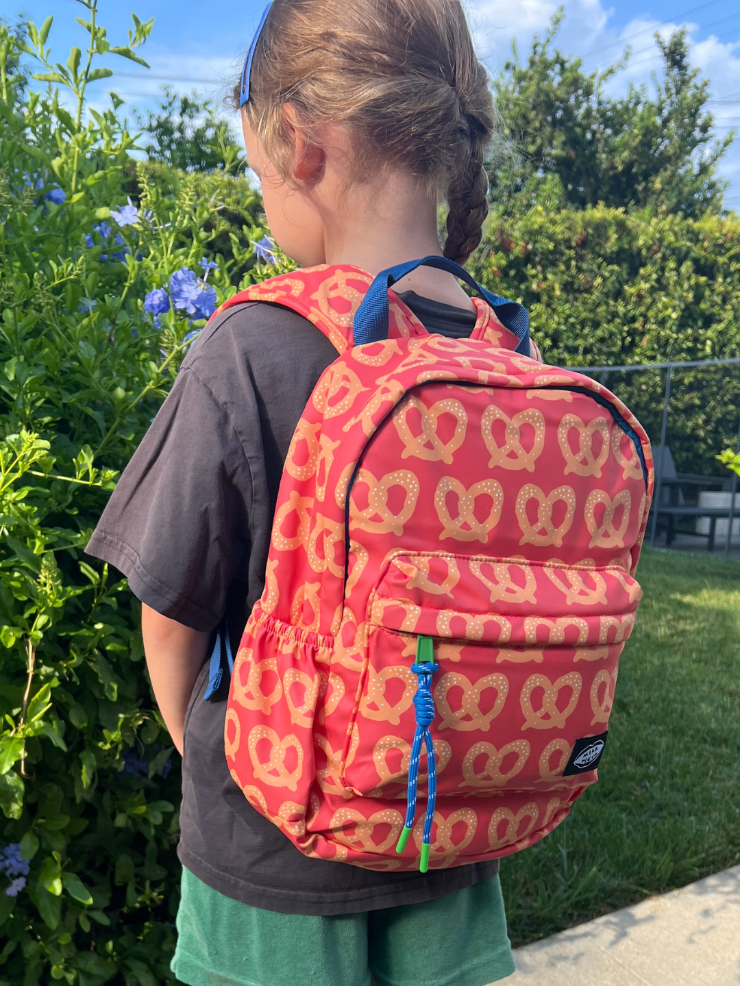 A child wearing a red backpack with a repeating pretzel pattern, photographed outdoors in a garden setting. 