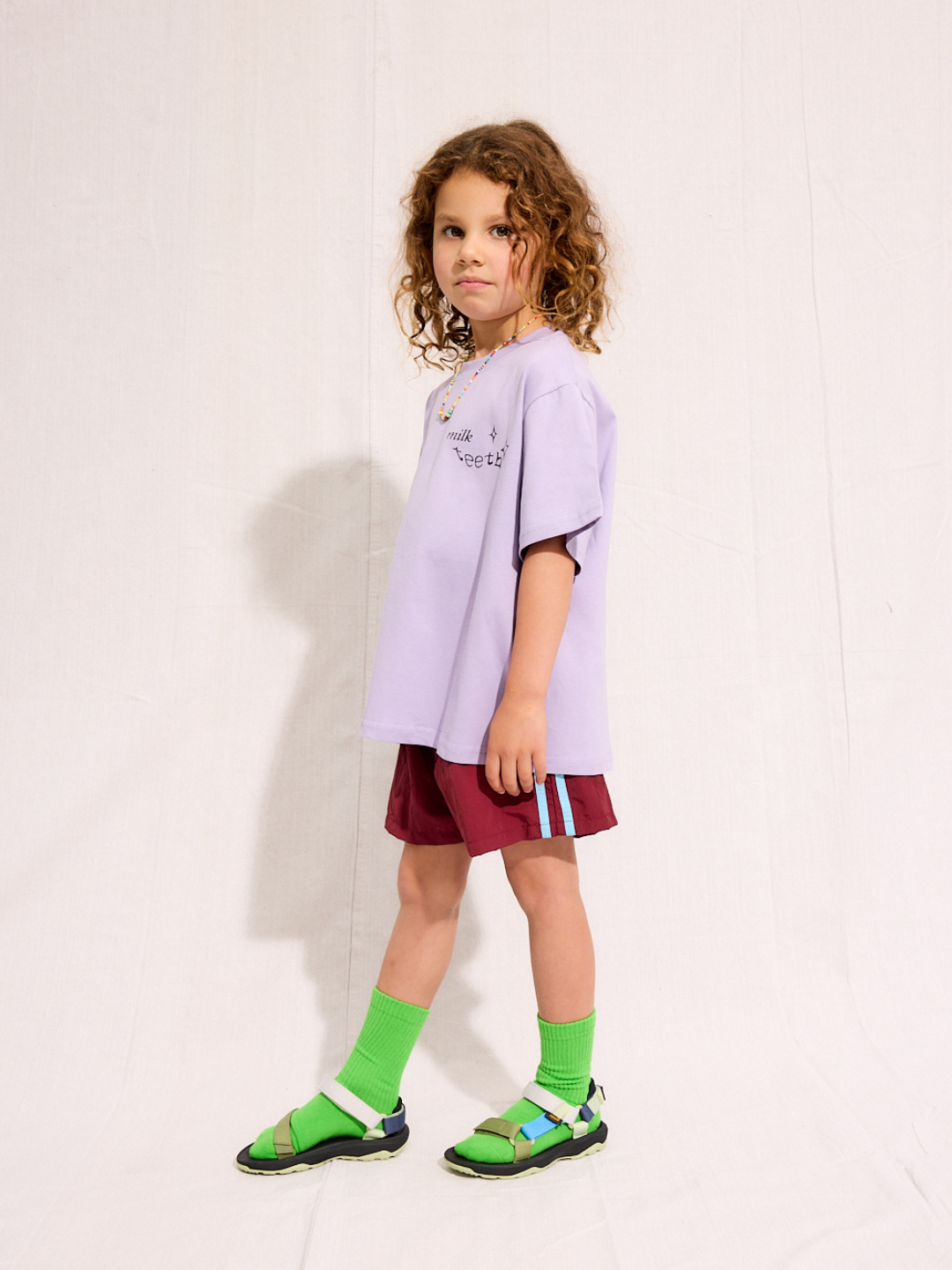 A child is wearing the purple Milk Teeth test with burgundy shorts and green socks and sandals