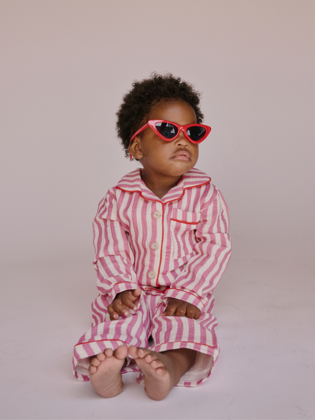 Child wearing a pink and white striped outfit with red cat eye sunglasses seated on a plain background