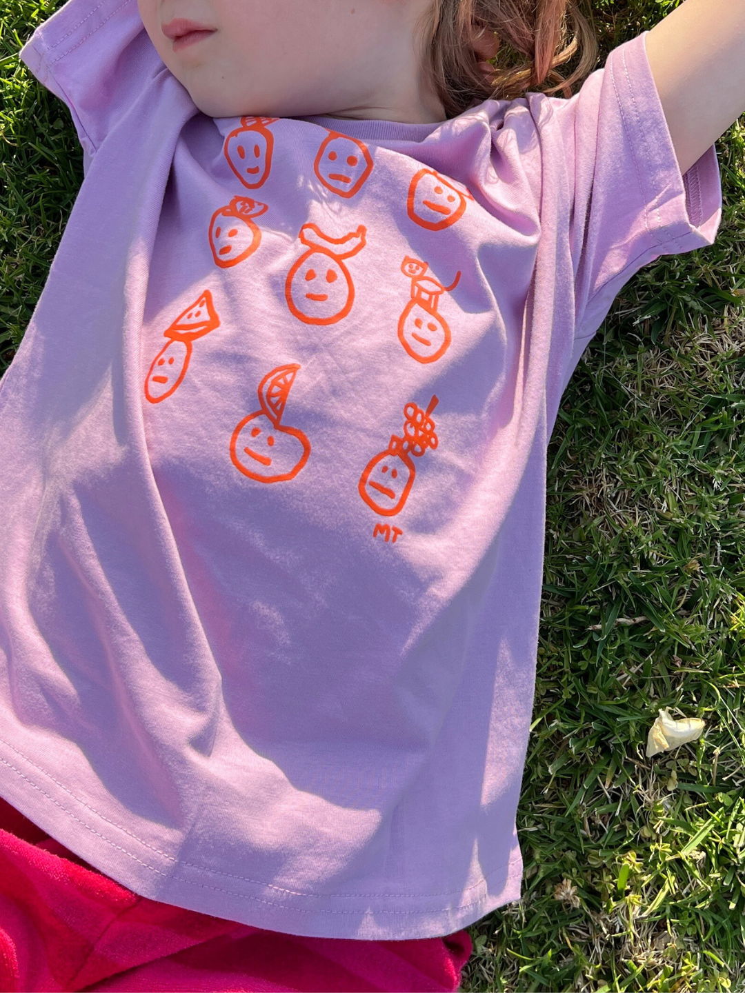 Violet | A child is laying on the grass wearing a Fruit Face tee in Violet with pink shirts. 