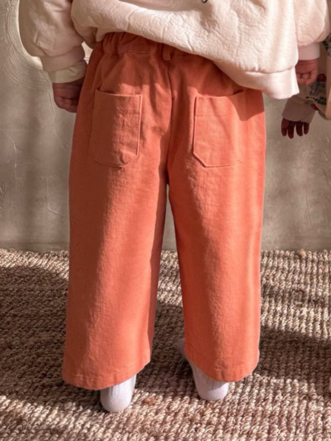 A child wearing Tomboy Pants in slouchy orange from a Korean kids brand and a cream sweater stands on a textured carpet.