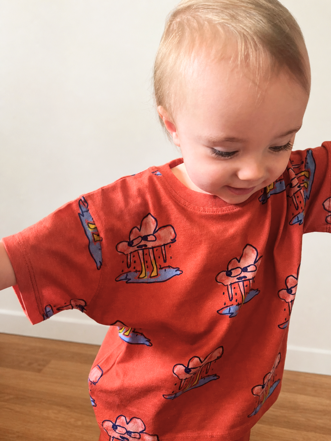 Red | A toddler indoors against a white wall, wearing the RAINY DAY SHORT SET,a children’s unisex matching t-shirt and shorts set in red, in soft organic cotton. It features a playful, repeating rain cloud character.