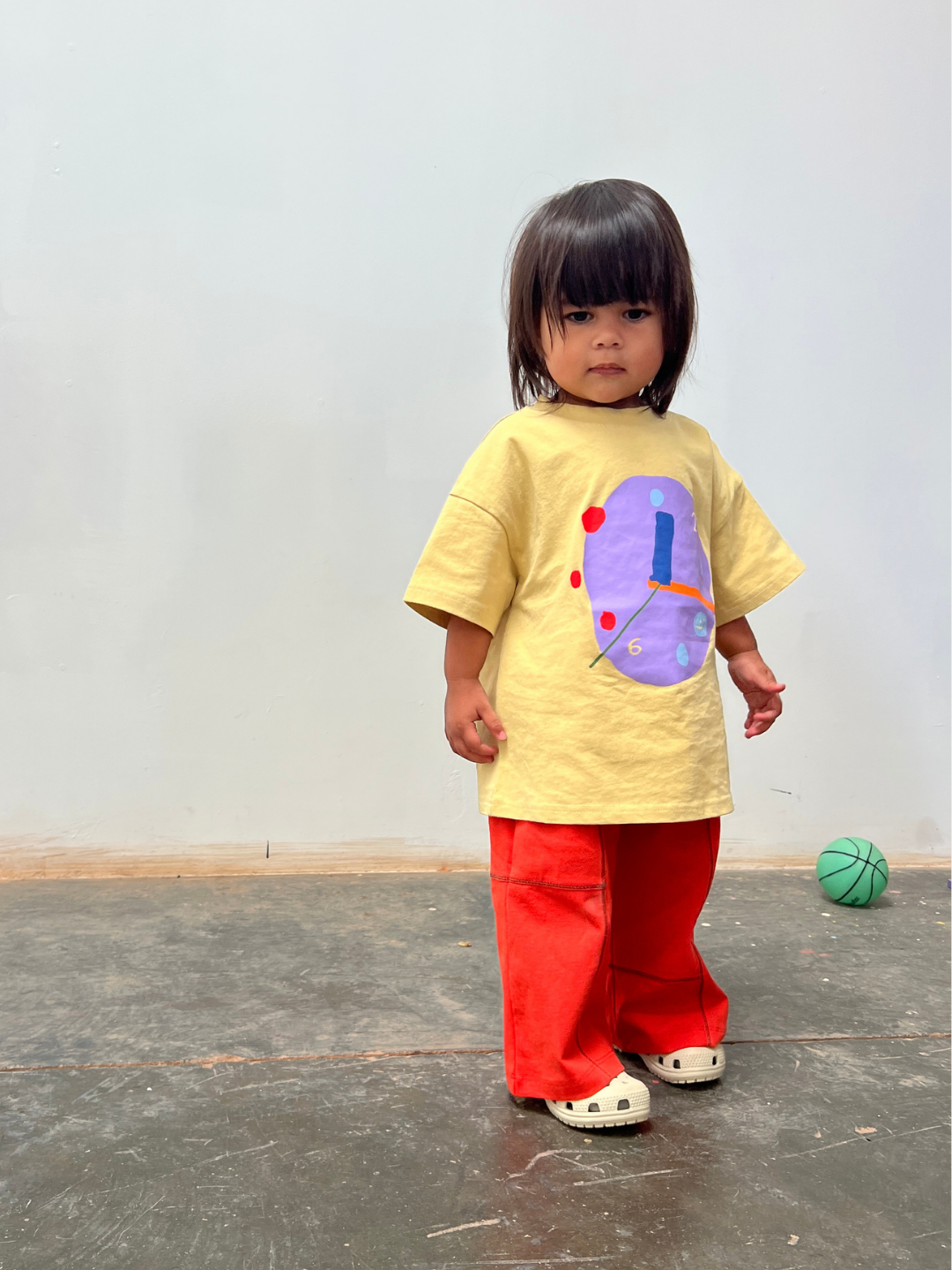 Yellow | Child wearing a yellow shirt with a colorful design and red pants standing on a gray floor.