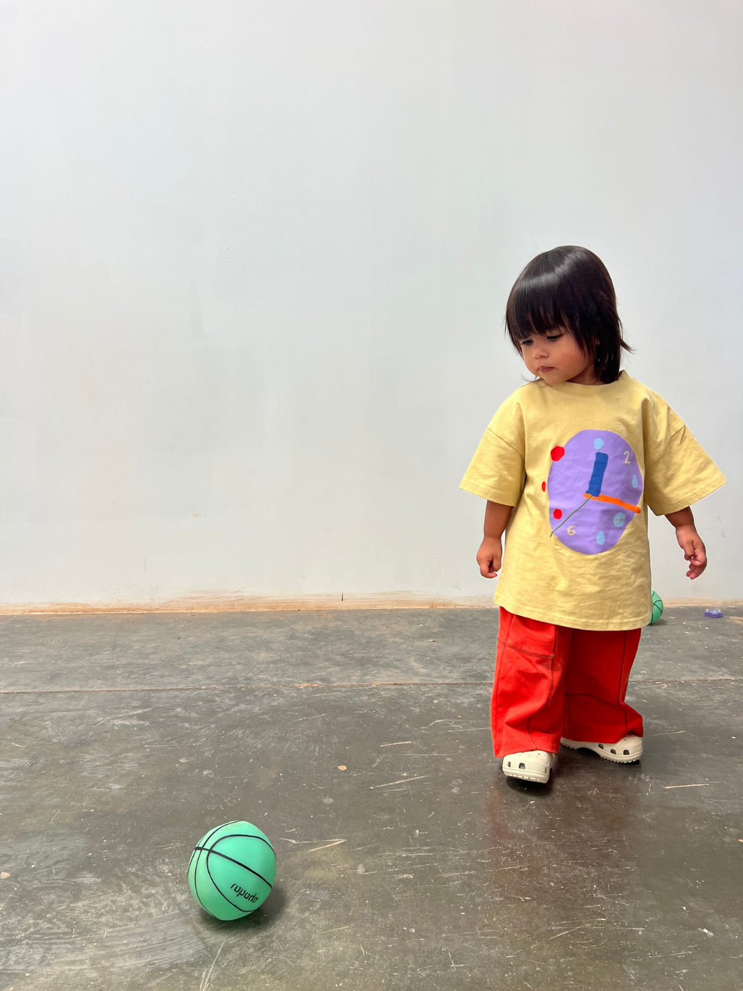Child in a yellow shirt with a colorful design and red pants standing next to a green ball on a gray floor.