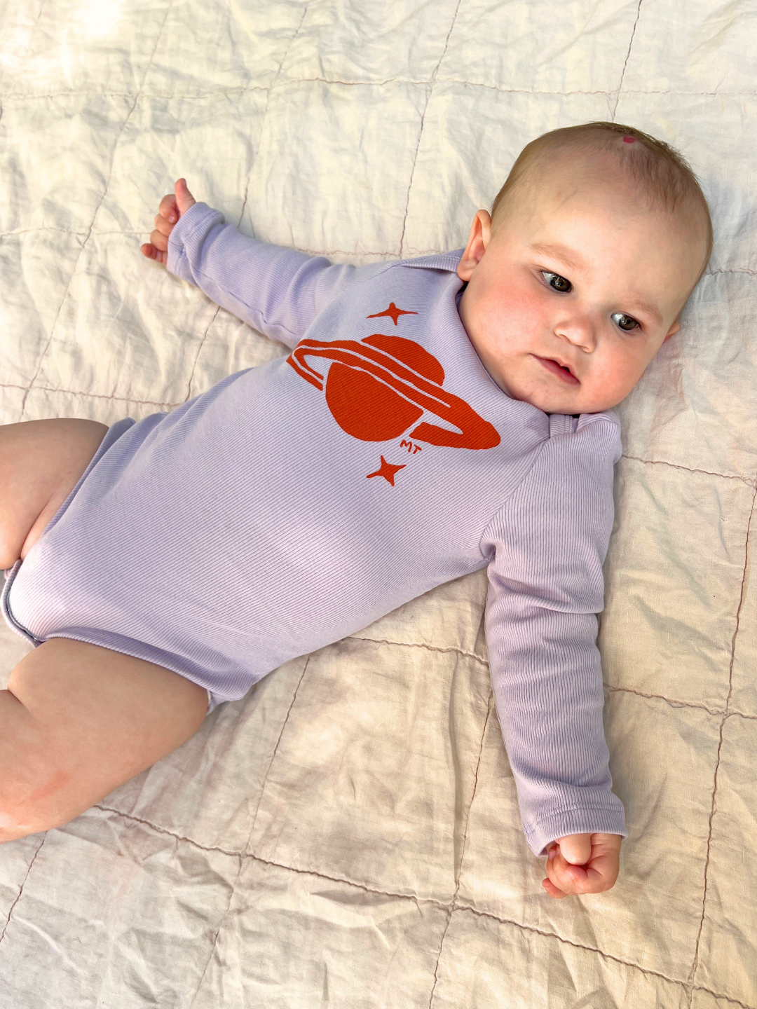 Lavender | Baby wearing a purple onesie with a red space-themed design on a light-colored blanket.