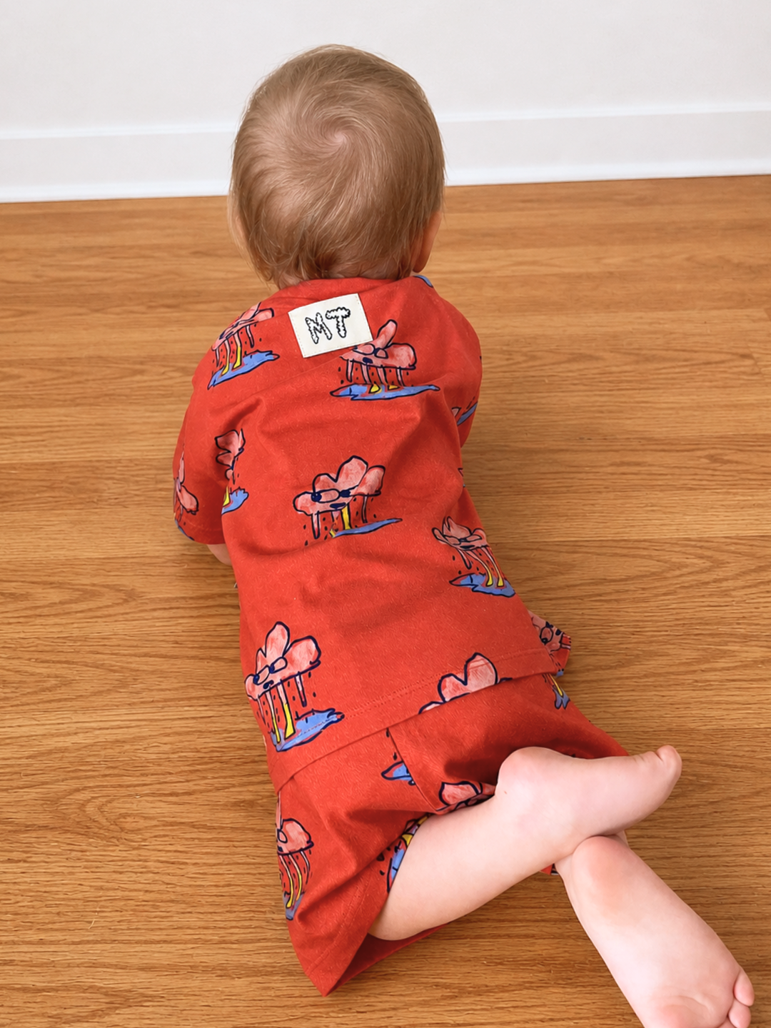 A toddler indoors lying on a wood floor, wearing the RAINY DAY SHORT SET,a children’s unisex matching t-shirt and shorts set in red cotton. It features a playful, repeating rain cloud character.