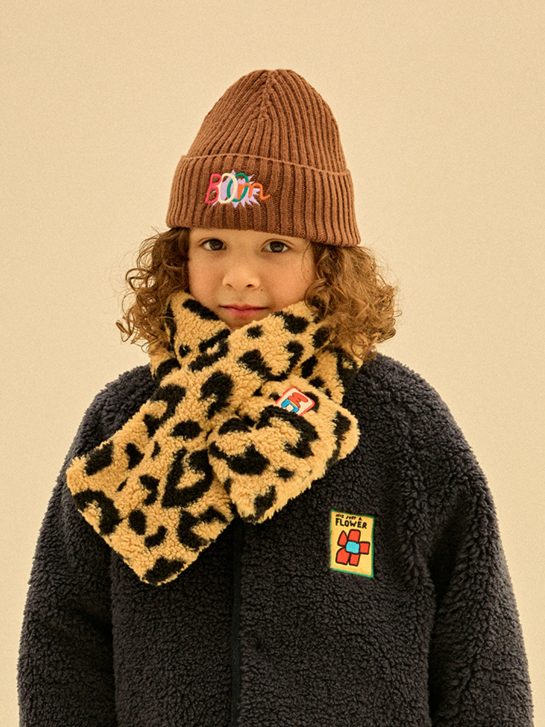 Child wearing a brown knit hat, the LEOPARD PATCH SCARF, and a dark fleece jacket with a small patch on the left side, standing against a plain background. This description could serve well for alt text in merchandising.