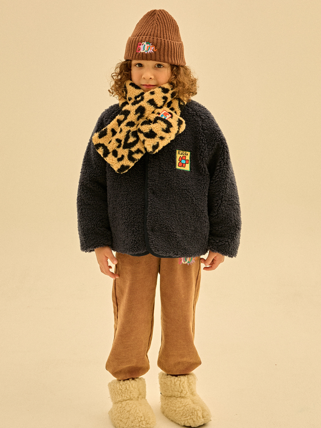 A child wearing a brown hat, the LEOPARD PATCH SCARF, a dark blue jacket, brown pants, and beige boots stands in front of a plain background. This description is ideal for alt text in merchandising catalogs.