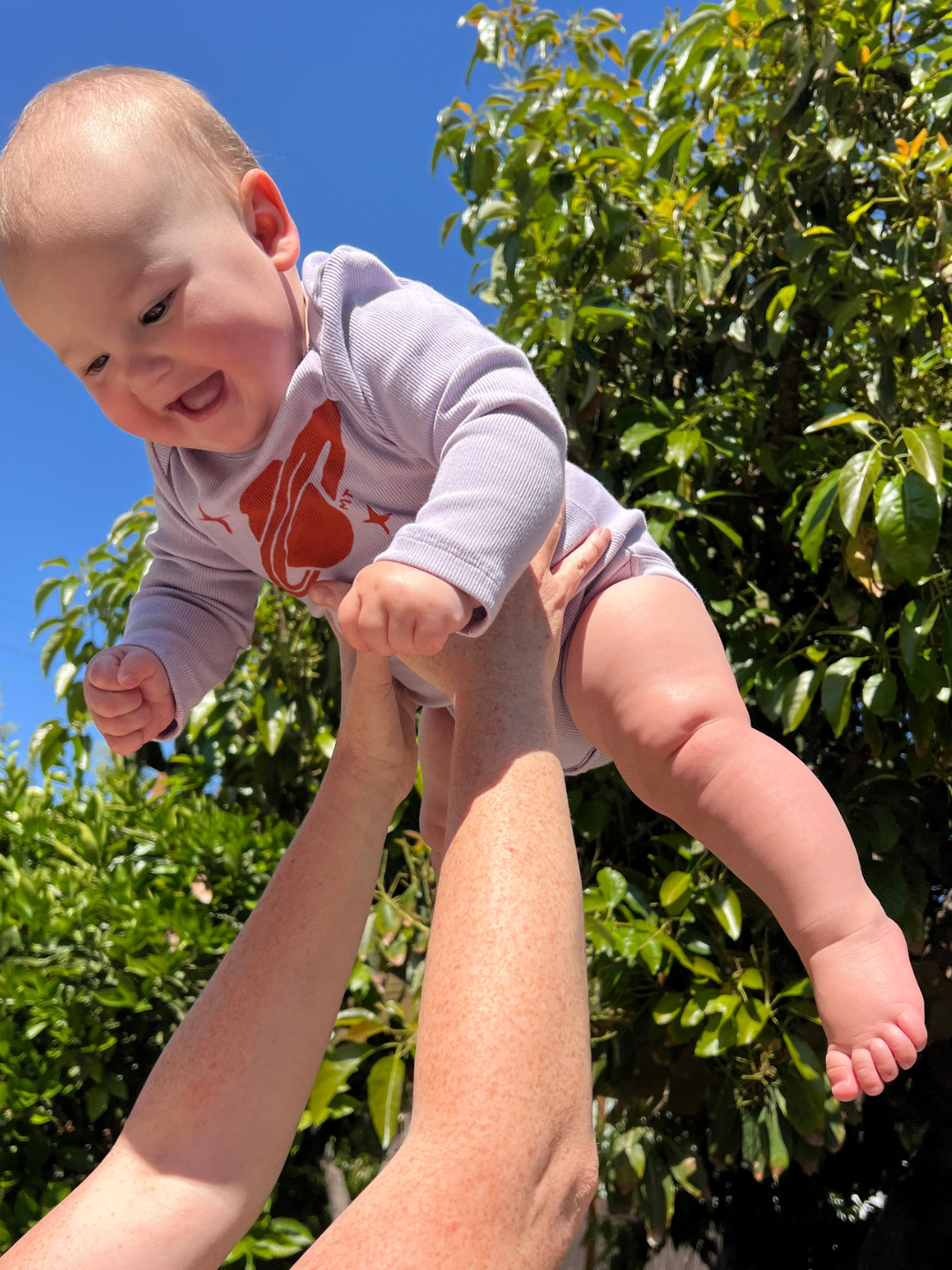 Baby in a light purple onesie with a red planet design being held up against a blue sky and green foliage.