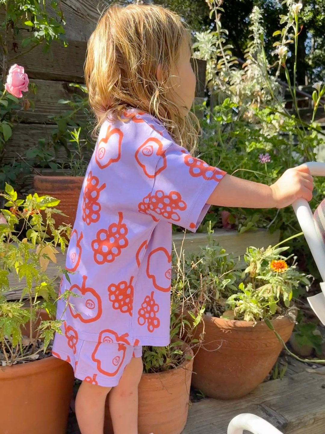 Child wearing the FRUIT STAND SHORT SET, a kids' matching outfit with a short-sleeve shirt and shorts, showcasing colorful apple and grape prints on a purple background, standing in a garden.