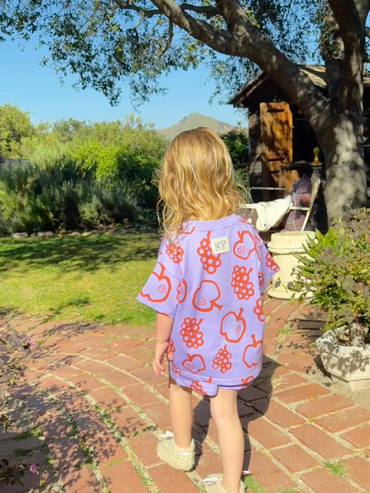Second image of The FRUIT STAND SHORT SET is a kids' matching outfit with a short-sleeve shirt and shorts, showcasing colorful apple and grape prints on a purple background, displayed on a plain white surface.