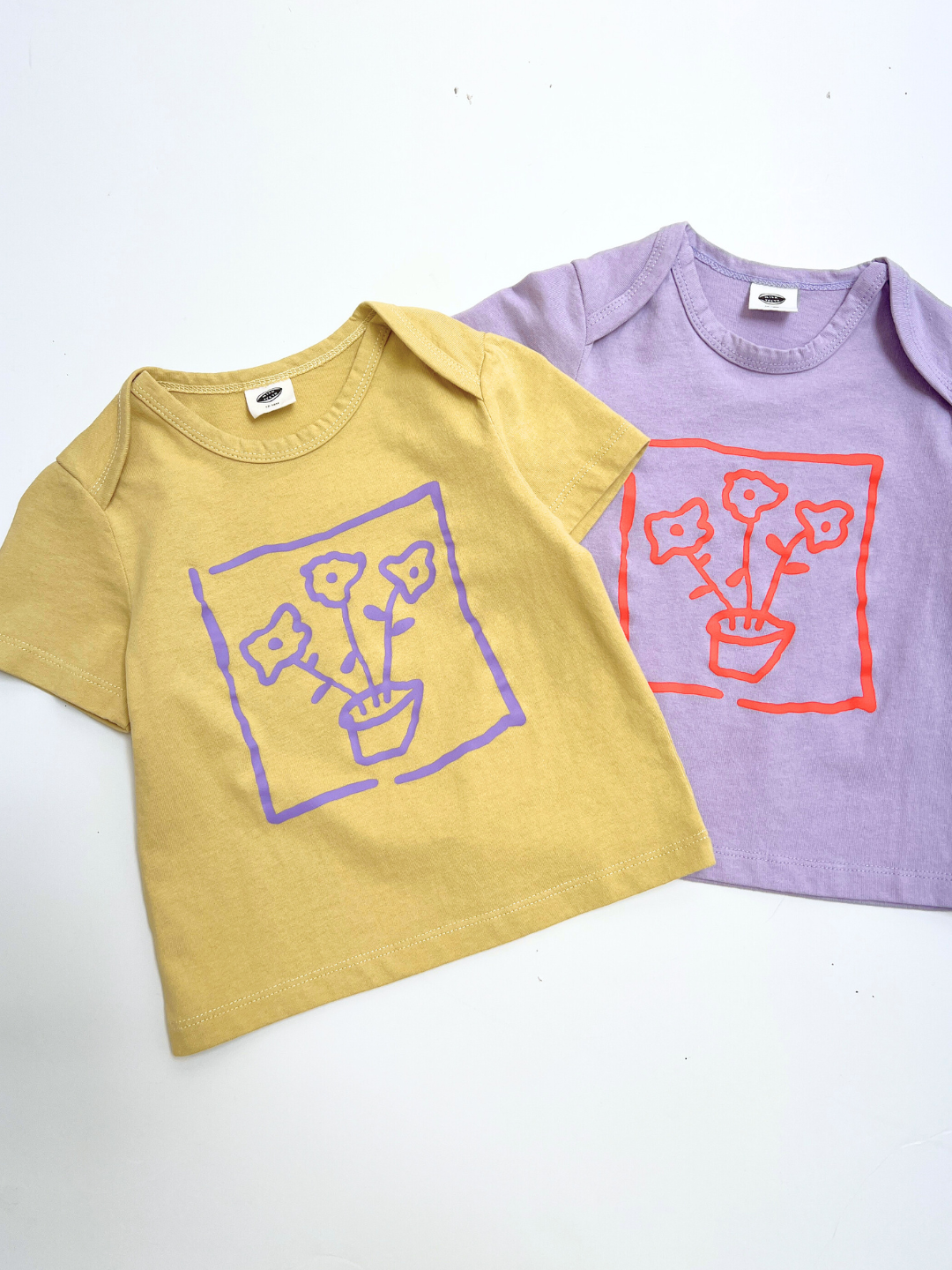 Yellow | Two FLOWER POT BABY TEEs for toddlers—one yellow with a purple flower pot graphic and one lavender with a red flower pot graphic—are laid flat on a white background.