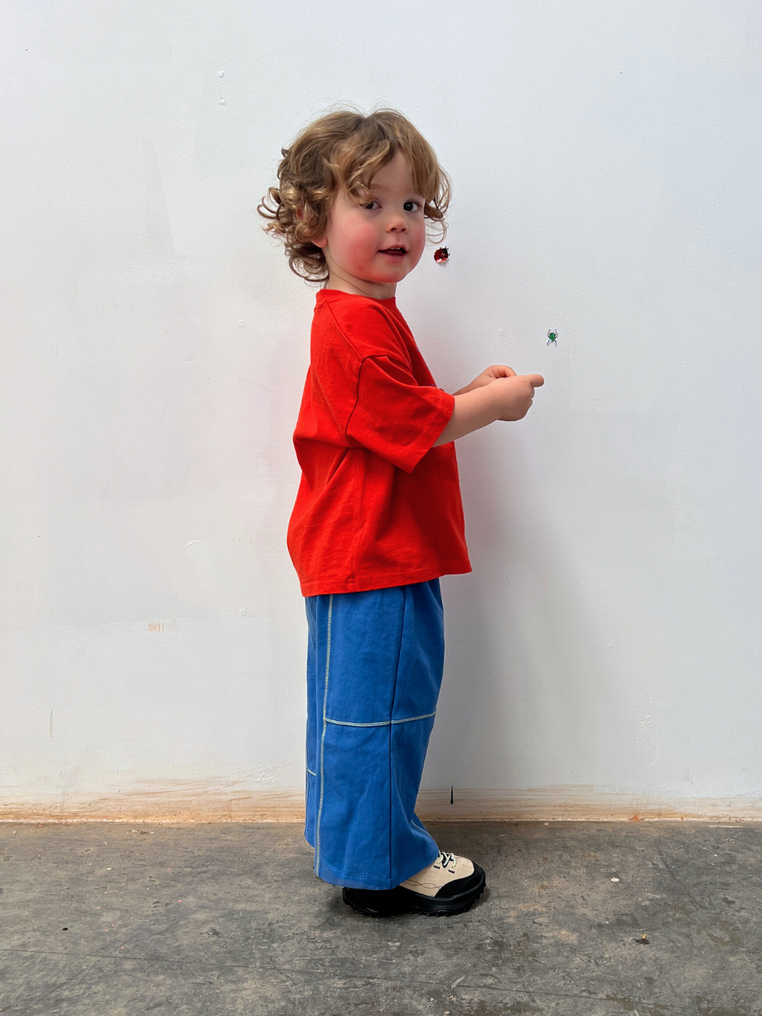 Blue | Child wearing a red shirt and blue pants standing against a white wall.