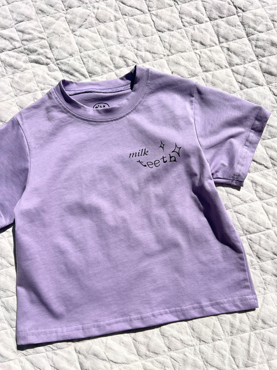 Purple | The MILK TEETH TEE, an artist-designed lavender short-sleeve shirt with black "milk teeth" text and star prints on the chest, is displayed on a quilted white surface.