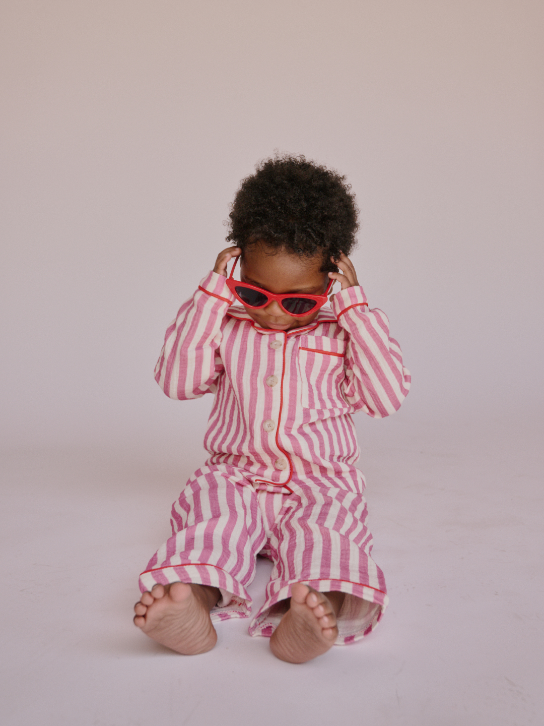Red | Child wearing a pink and white striped outfit with red cat eye sunglasses seated on a plain background