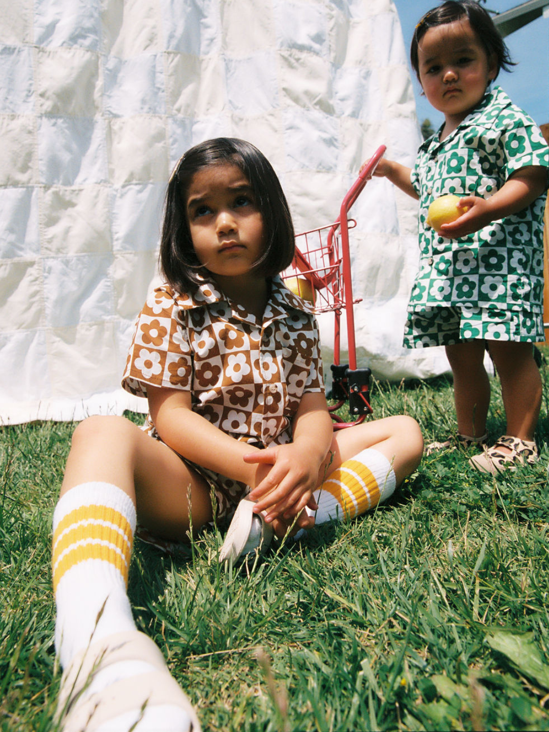 Two children on the grass wearing kids' shirt and shorts sets, one in a checkerboard pattern of terracotta and white flowers, the other with green and white flowers