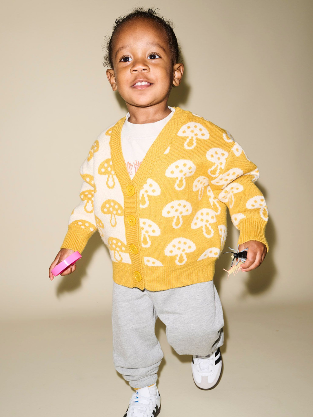 A child in a SHROOM CARDIGAN, grey pants, and white sneakers holds small objects while smiling and walking, creating a heartwarming scene that defies any detailed description.