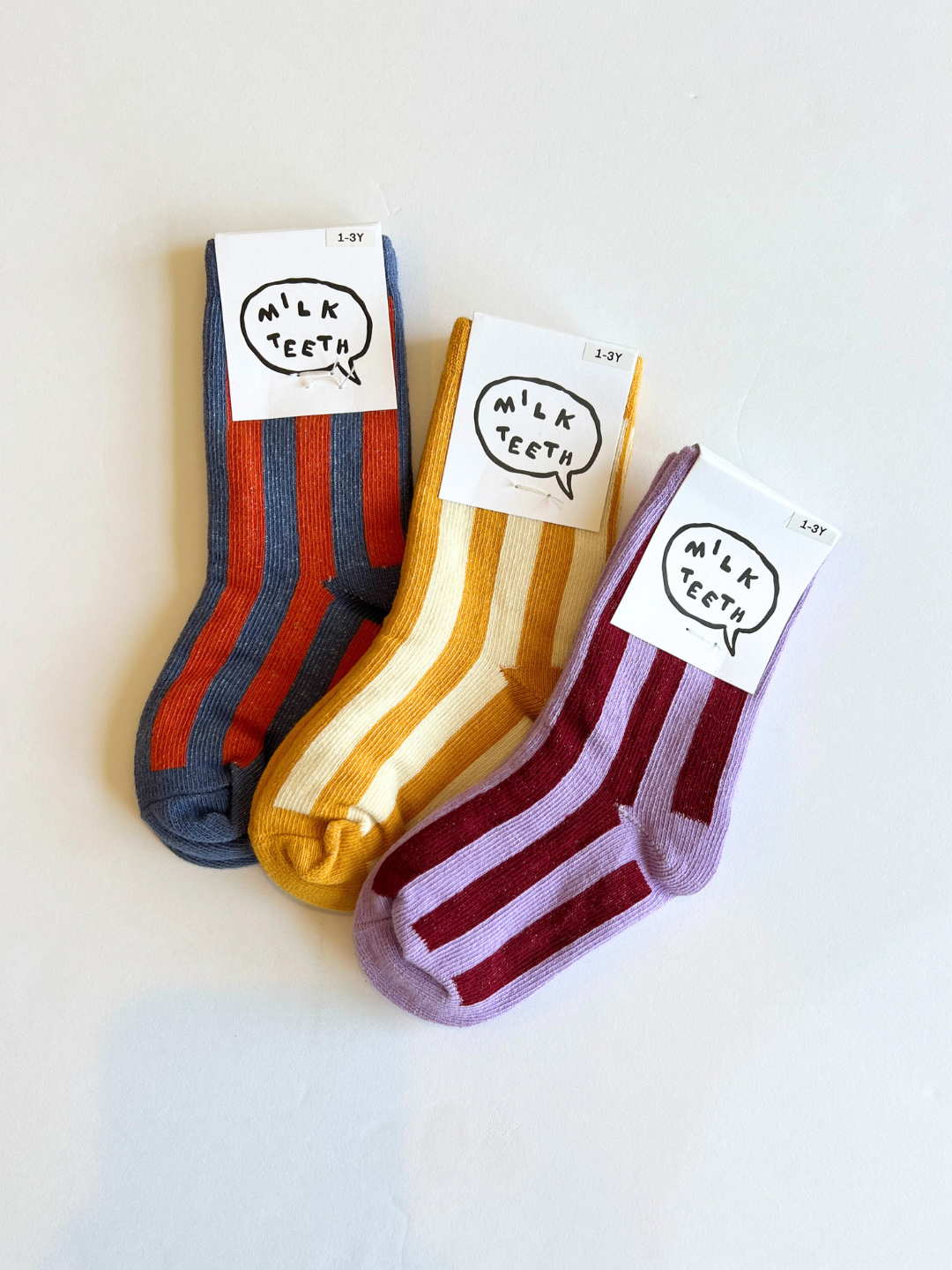 Slate | Three pairs of children's STRIPE SOCK are displayed with labels reading "Milk Teeth." Available in blue and red, yellow and white, and purple and red, these comfy socks are made from stretchy cotton, making them perfect for little feet.