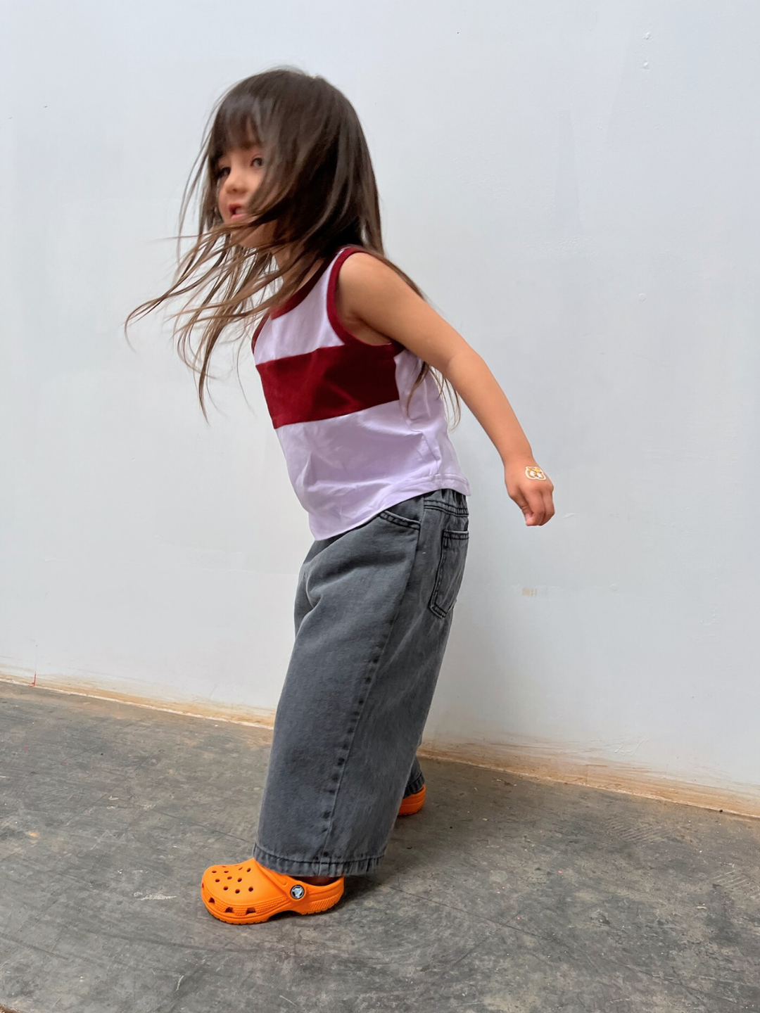 Purple/Maroon | A young child stands indoors against a plain light wall, wearing the TRACK TANK—a colorblocked cotton tank top—paired with wide-leg gray relaxed-fit jeans and orange clogs.