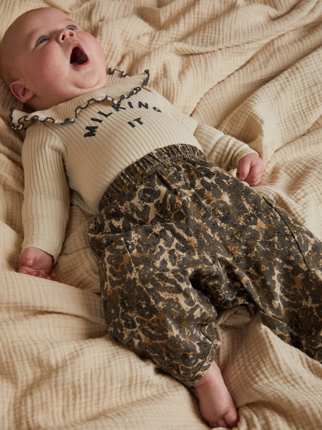 Baby wearing a sweater with text and leopard print pants on a beige blanket