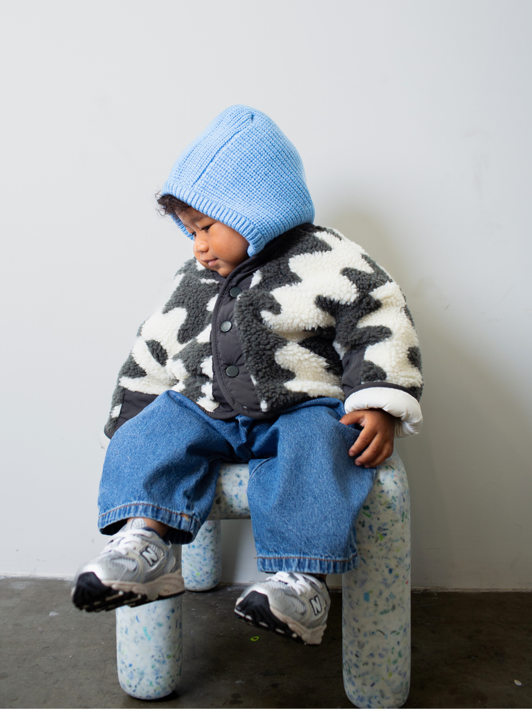 Sky Blue | Child sitting on a small stool wearing a patterned jacket, sky blue hood and blue jeans against a plain background