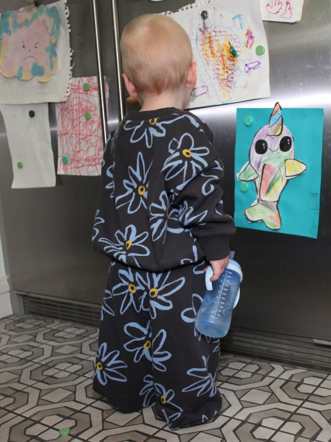 A young child wears the CHARLIE SWEAT SET in a painted flower print French Terry fabric, standing on a patterned floor, facing a fridge covered in colorful children's drawings, and holding a blue sippy cup.