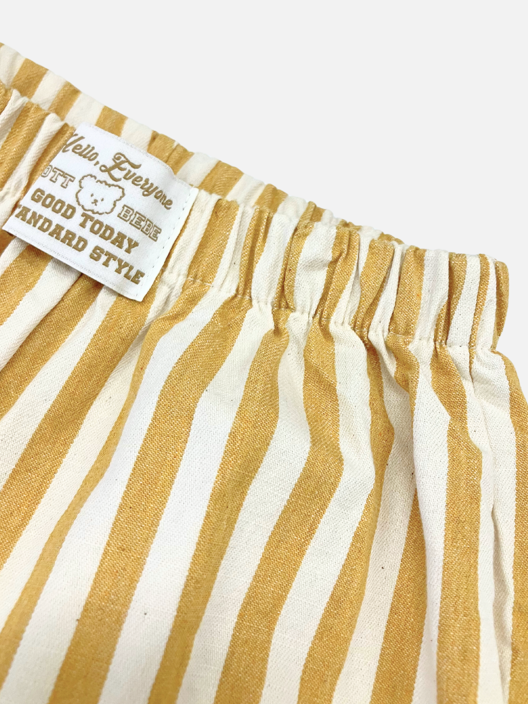 Mustard | Close-up of the PONY STRIPE PANTS, featuring yellow and white vertical stripes in striped cotton twill and an elasticated waistband. A sewn-on label reads, "Hello Everyone, GOOD TODAY, STANDARD STYLE" with a bear graphic.