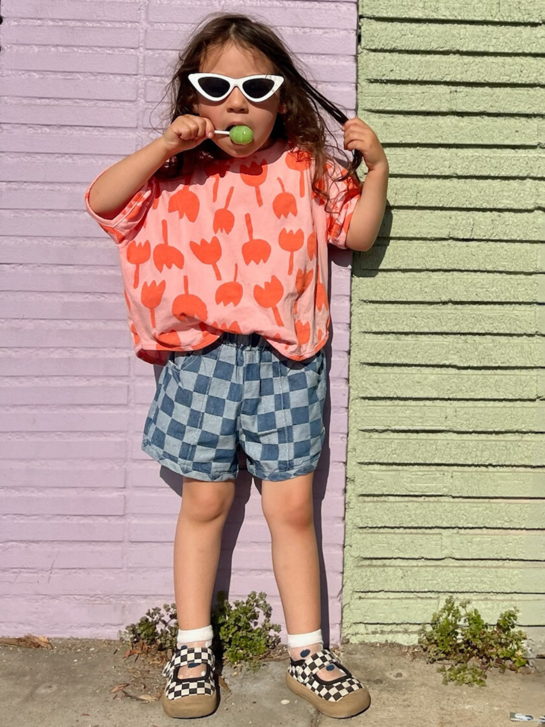 A young girl stands against a wall with purple and green sections. She wears white sunglasses, an orange and pink tulip-patterned top, vintage CHECKMATE SHORTS, checkerboard slip-on shoes, and holds a green lollipop in her mouth.