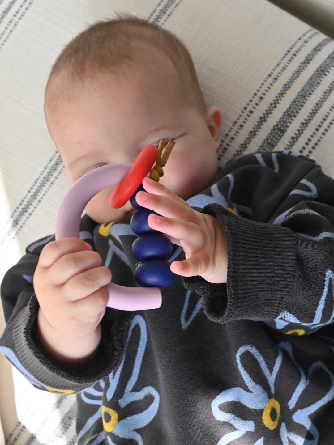 A baby lies on a striped surface, holding the MT X JM ARCH RING TEETHER with both hands while wearing a dark sweater with large floral patterns.