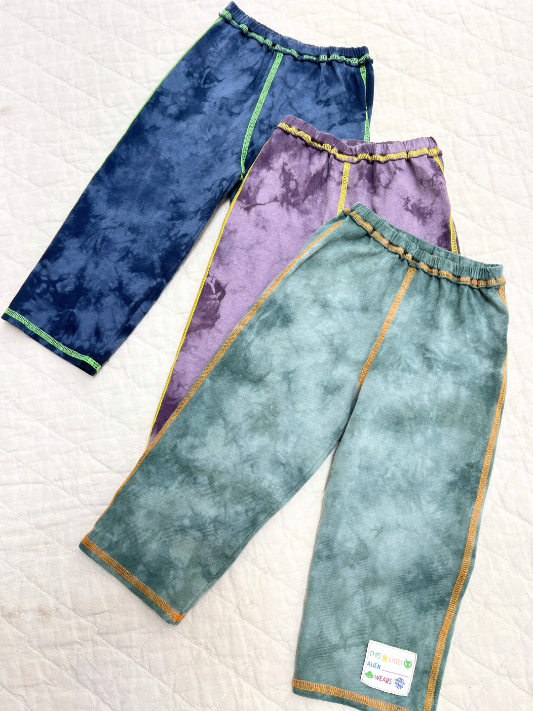Sage | Three pairs of COSMO TIE DYE PANT in blue, purple, and green with colored stitching are laid out on a light quilted surface.