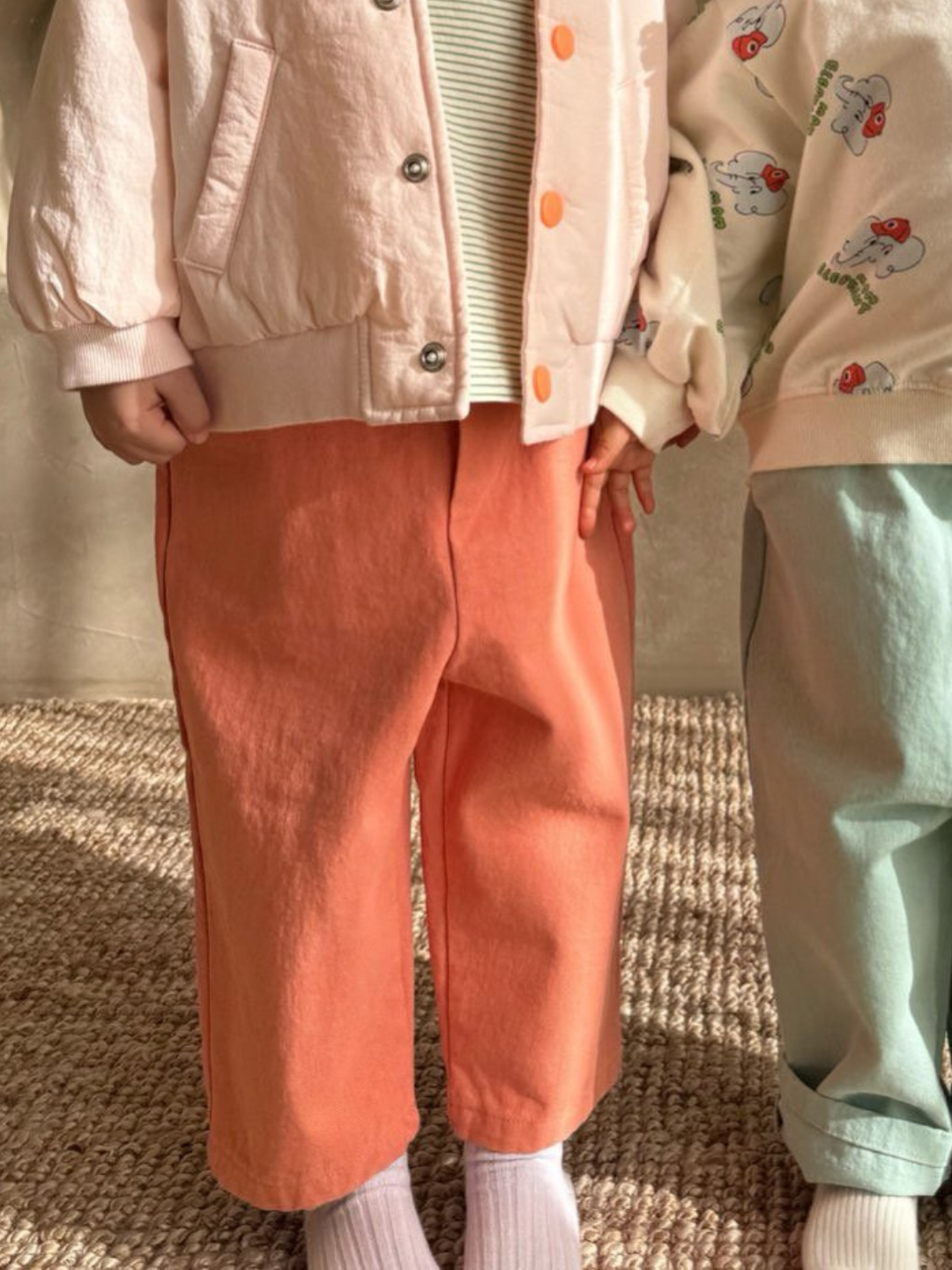 Two children showcase a Korean kids' brand in pastel outfits. One wears cotton twill coral Tomboy Pants and a pink jacket, while the other is in teal pants and a printed top, both made from cotton twill. They stand on a carpeted floor.