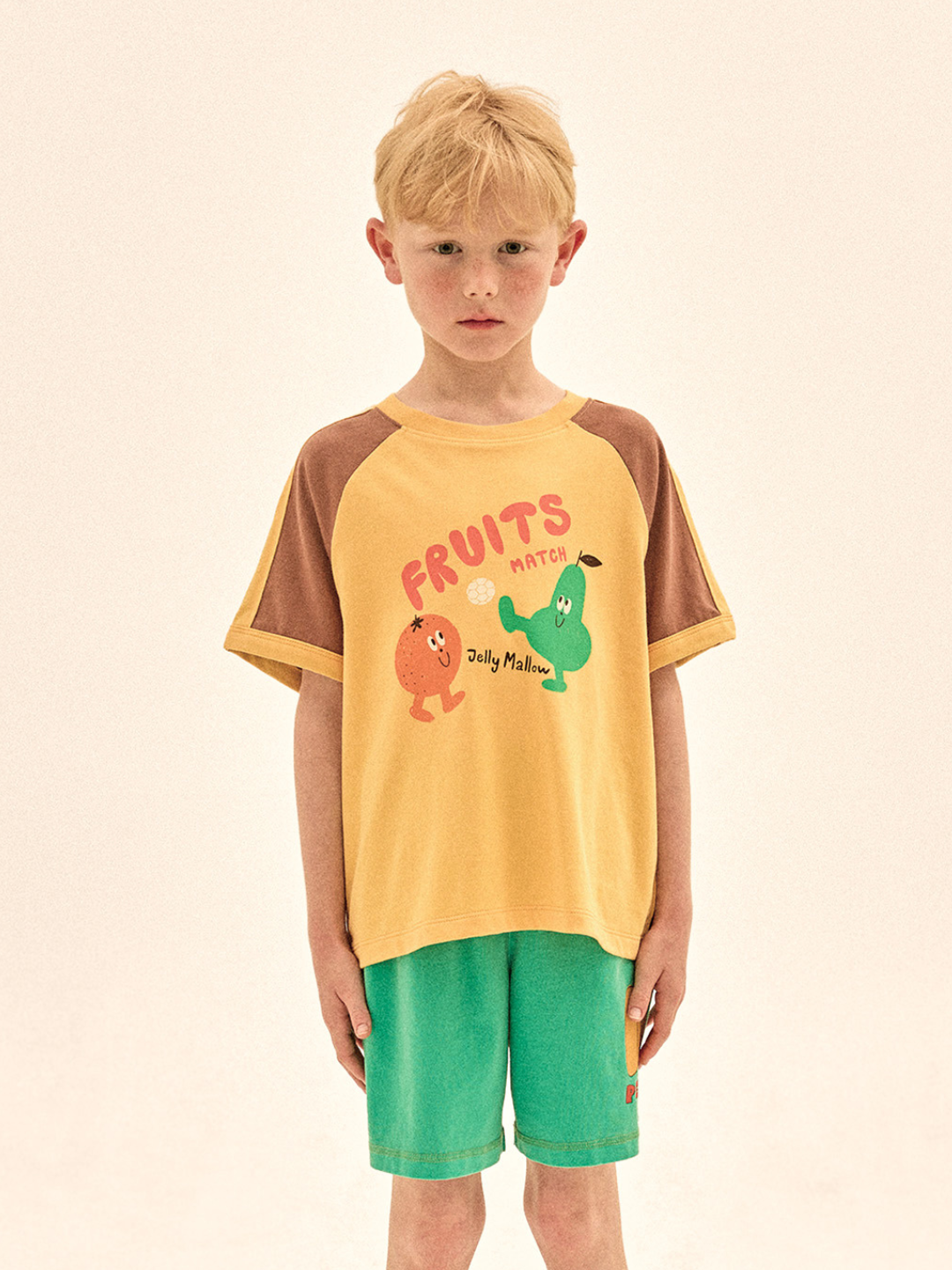 Child wearing a yellow and brown shirt with cartoon characters and green shorts on a beige background