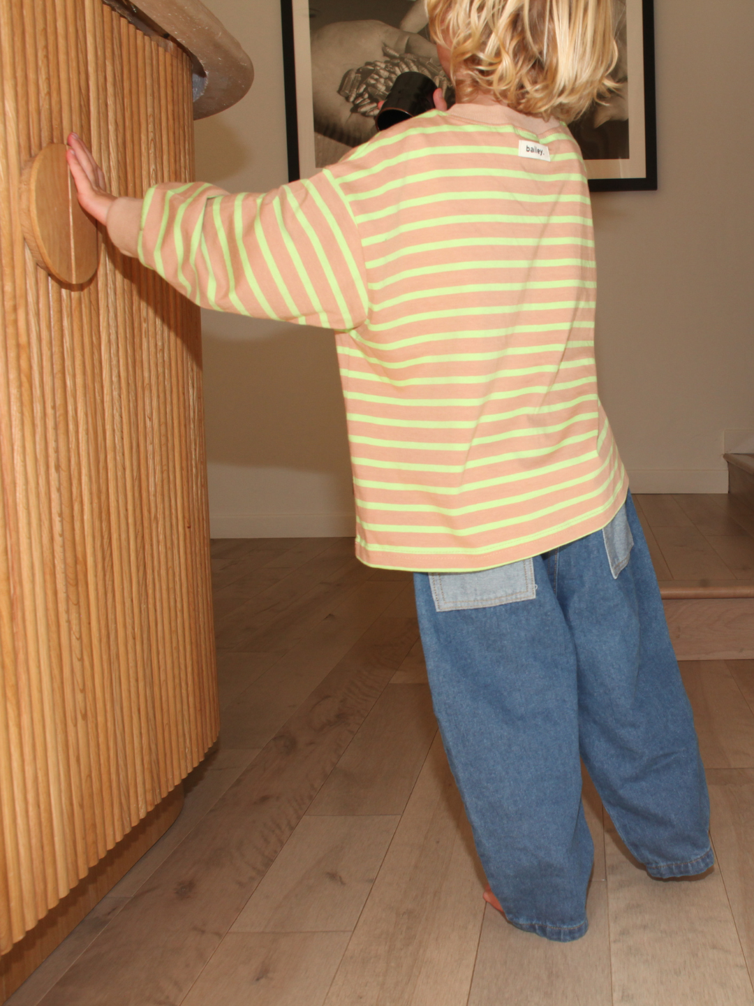 A child wearing a striped shirt and baggy denim pants standing on a wooden floor.