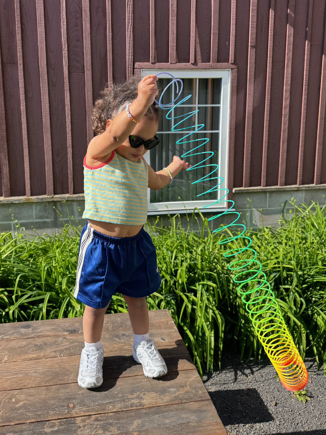 Child playing with a colorful slinky outdoors on a wooden deck, wearing black cats eye sunglasses, a green top and blue shorts