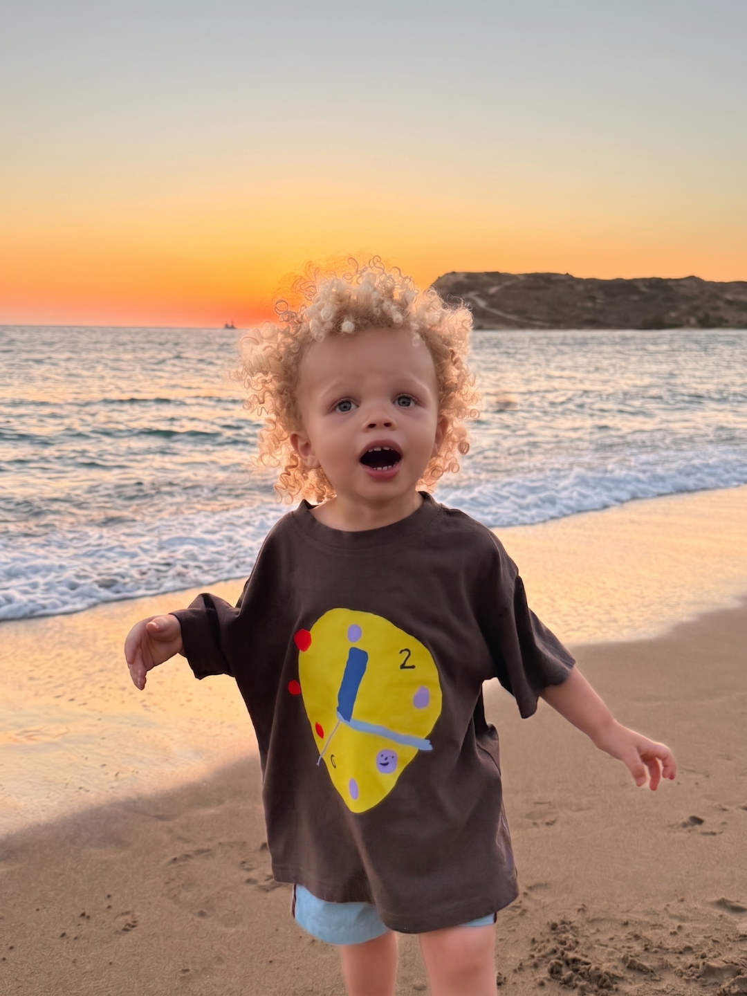 A toddler with curly blonde hair stands on a sandy beach at sunset wearing an oversized dark brown t-shirt with a large yellow clock graphic on the front and light blue shorts.