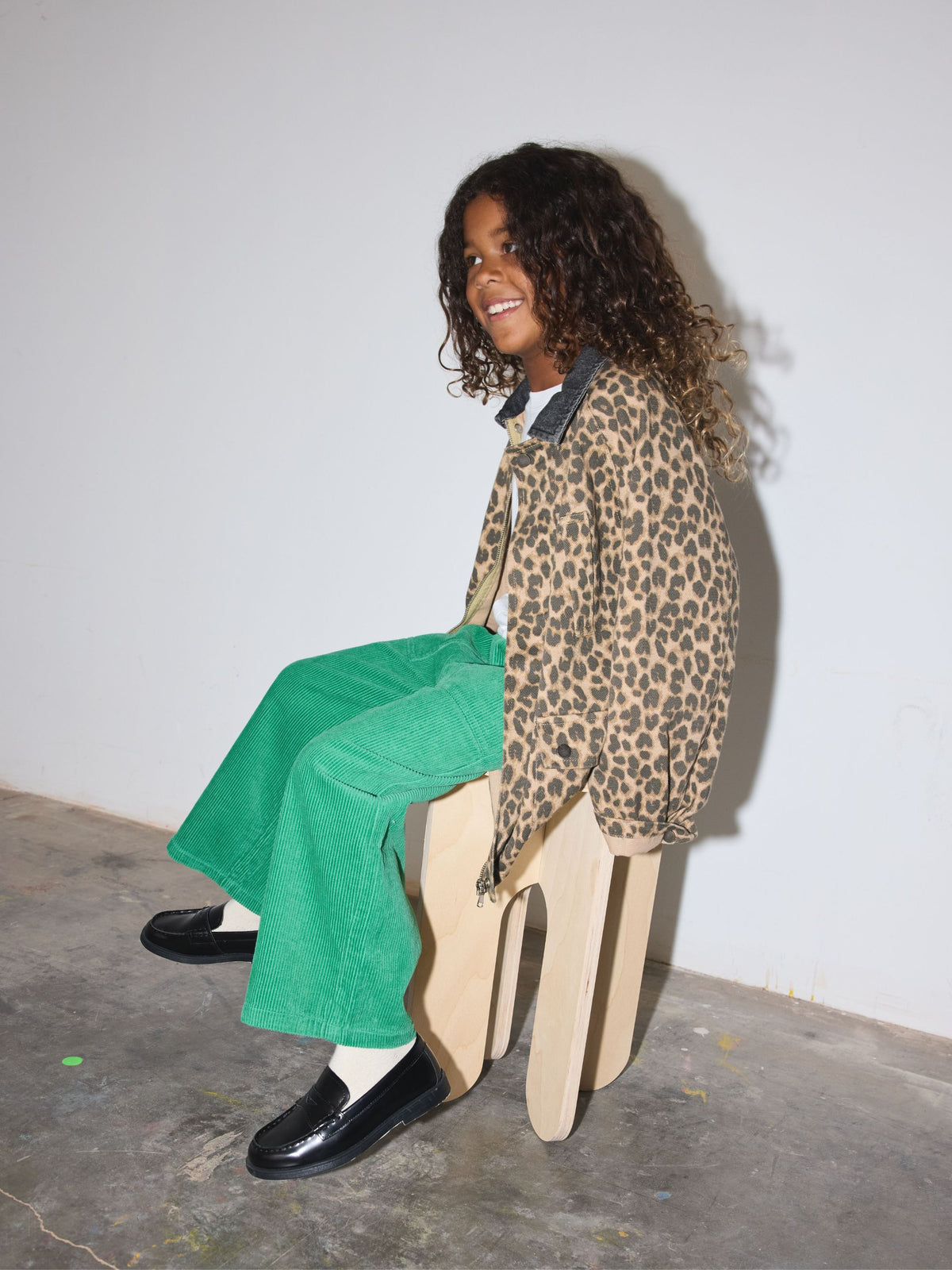 A smiling child sits on a light wood stool wearing a leopard print jacket, bright green corduroy trousers, a white t-shirt, and black loafers against a plain white wall.