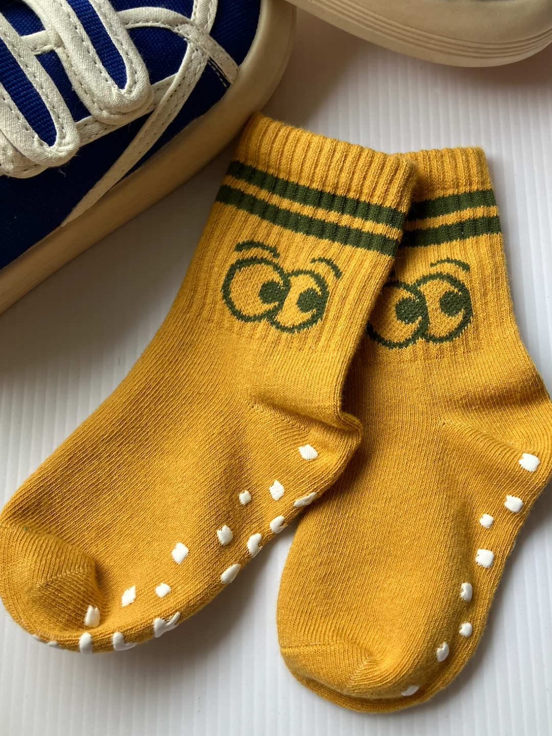 The RIBBED GRIP SOCK SET includes a pair of yellow, anti-skid grip socks with a green striped design featuring eyes, perfect for little ones when placed next to a pair of blue and white sneakers.