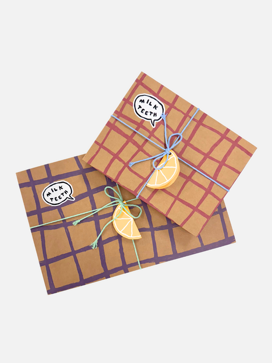 Second image of Two GIFT WRAP boxes featuring a purple grid pattern, embellished with lemon slice decorations, blue string, and a "Milk Teeth" speech bubble sticker, arranged on a monochrome patterned surface. Inside each box is a charming gift card and specialty tissue paper for an extra touch of elegance.