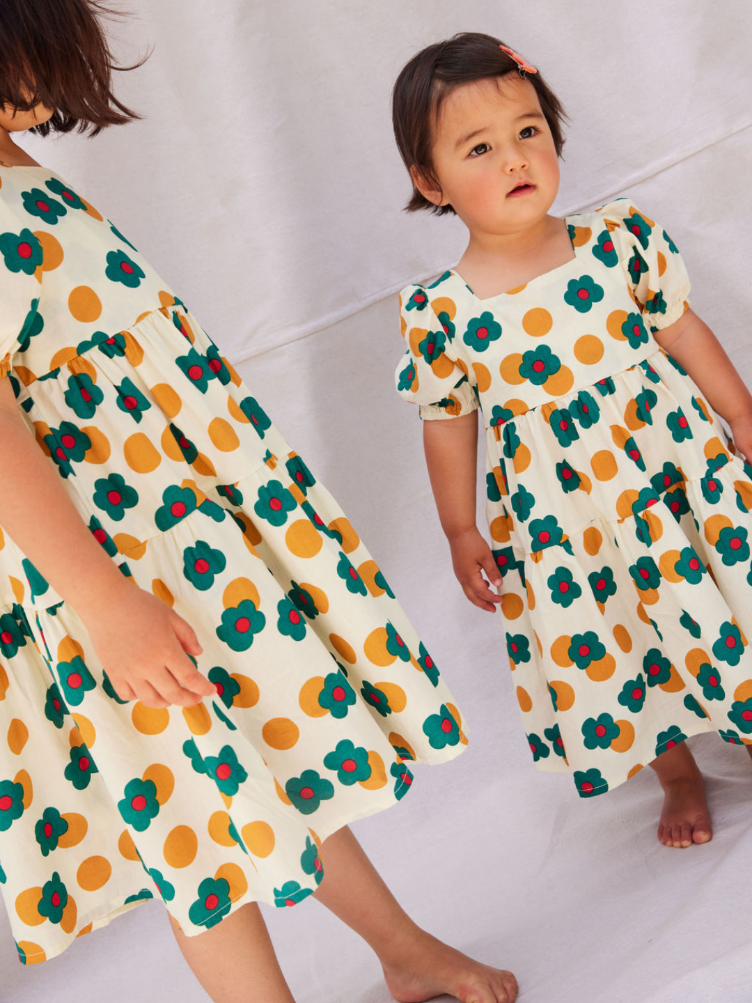 Two children wearing a kids' tiered, puff-sleeved dress in a pattern of green and red flowers and ochre dots on a white background