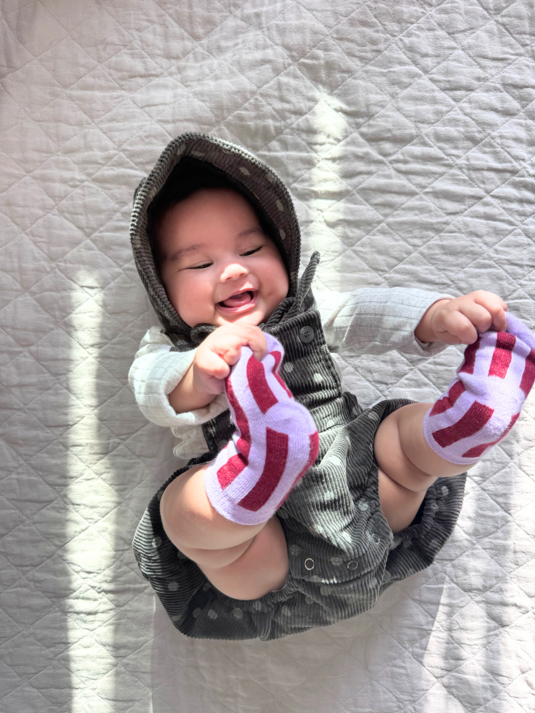 Violet | A smiling bag wears the STRIPE SOCKS: A pair of light violet, stretchy cotton socks featuring dark red vertical stripes. They wear a grey corduroy romper and matching bonnet, printed all over with polka dots. They are lying on a beige quilt and holding their toes.