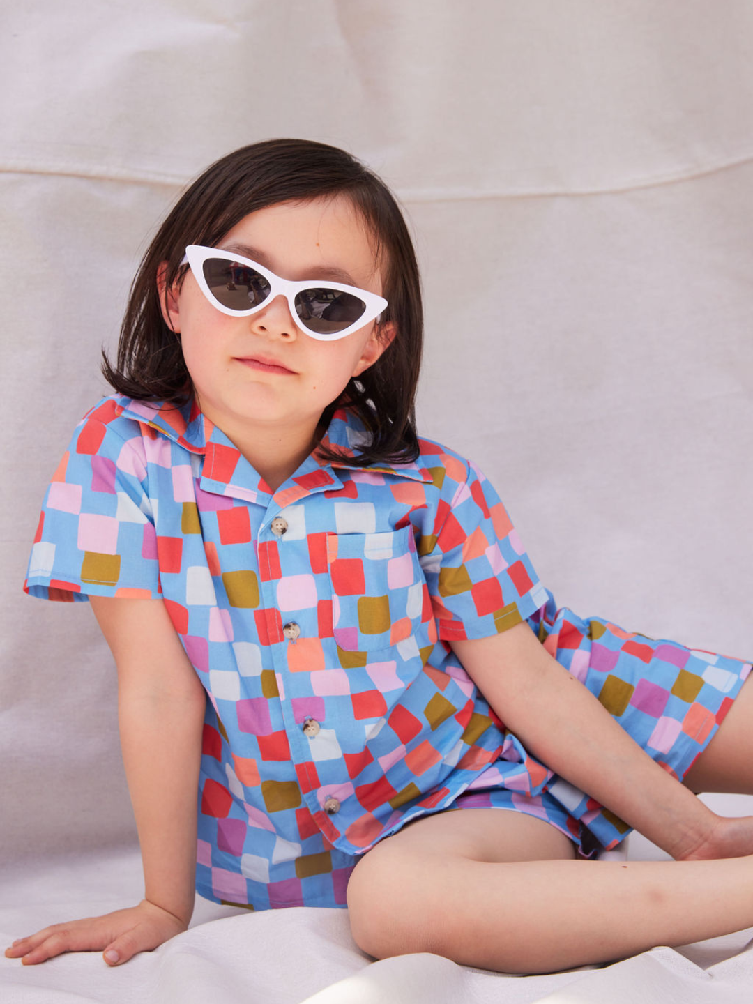 White | A child wearing the white Stingray kids Sunglasses. She is seated on a beige cloth backdrop and wears a blue shorts set patterned with multicolored squares.