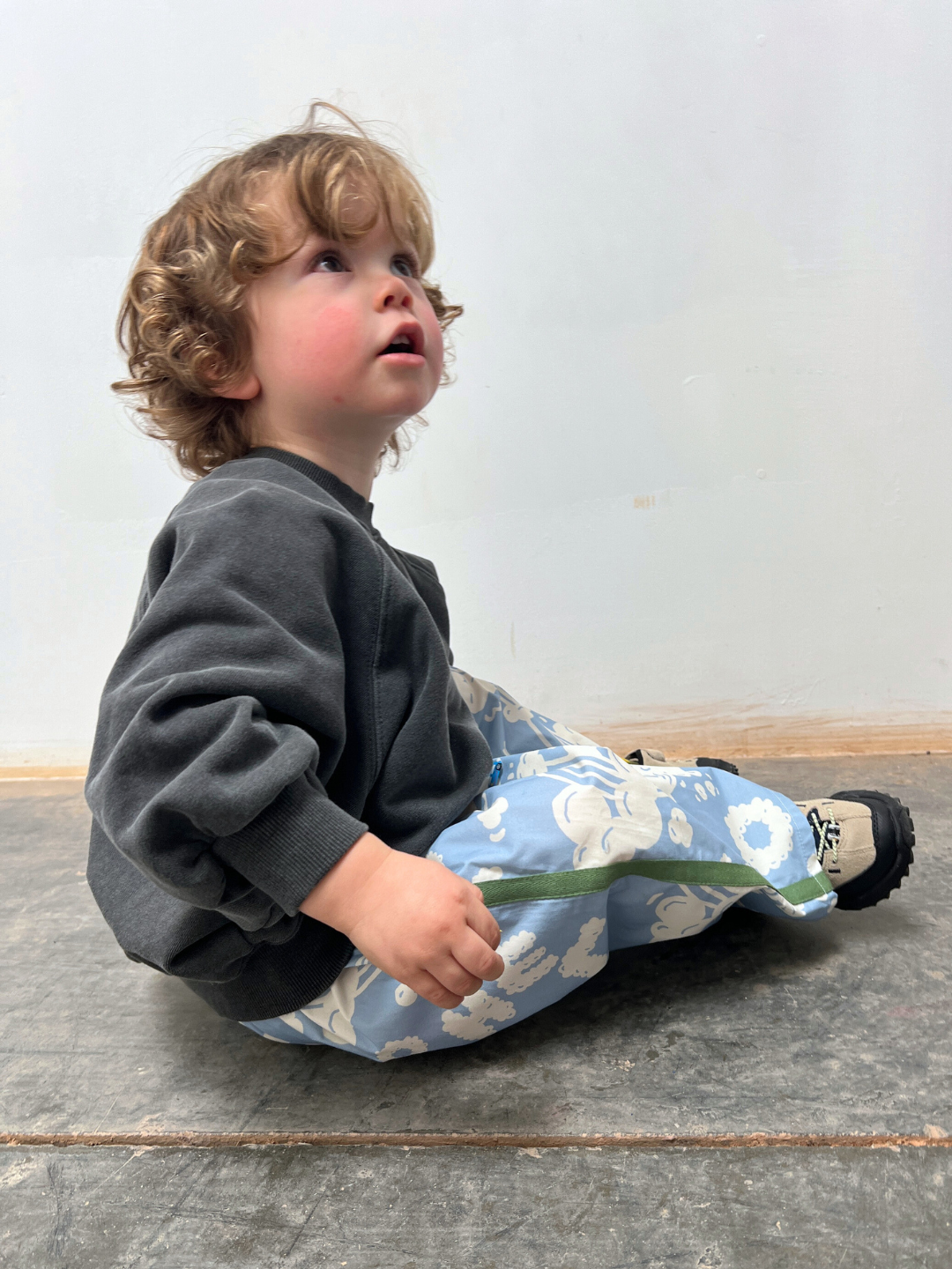 Child sitting on a wooden floor wearing a gray sweatshirt and floral-patterned pants.