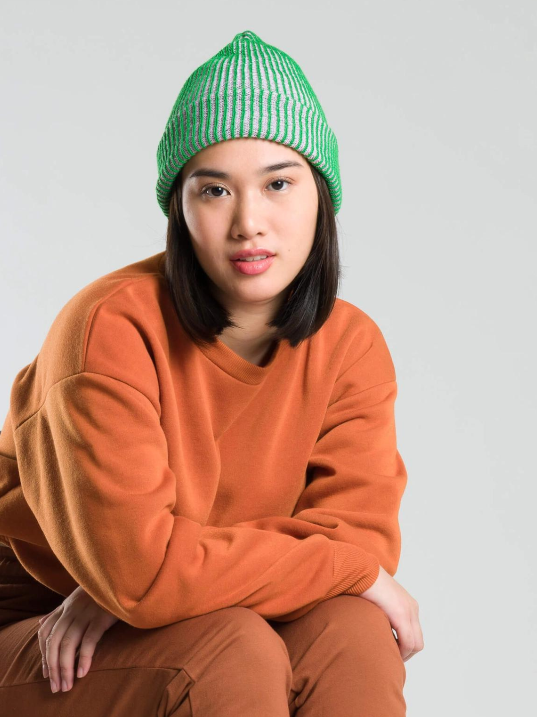Kelly/Lilac | A woman wearing a kelly green ribbed beanie for big kids and adults, with a ribbed cuff. The ribbed texture reveals a second yarn color that's light purple. She is wearing a rust orange sweatshirt and pants, and is seated in front of a grey wall.