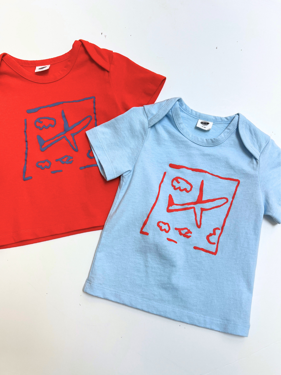 Two AIRPLANE BABY TEE shirts for toddlers—one red, one light blue—both with a cute airplane and cloud print. Ideal playful design for little ones who love planes!.