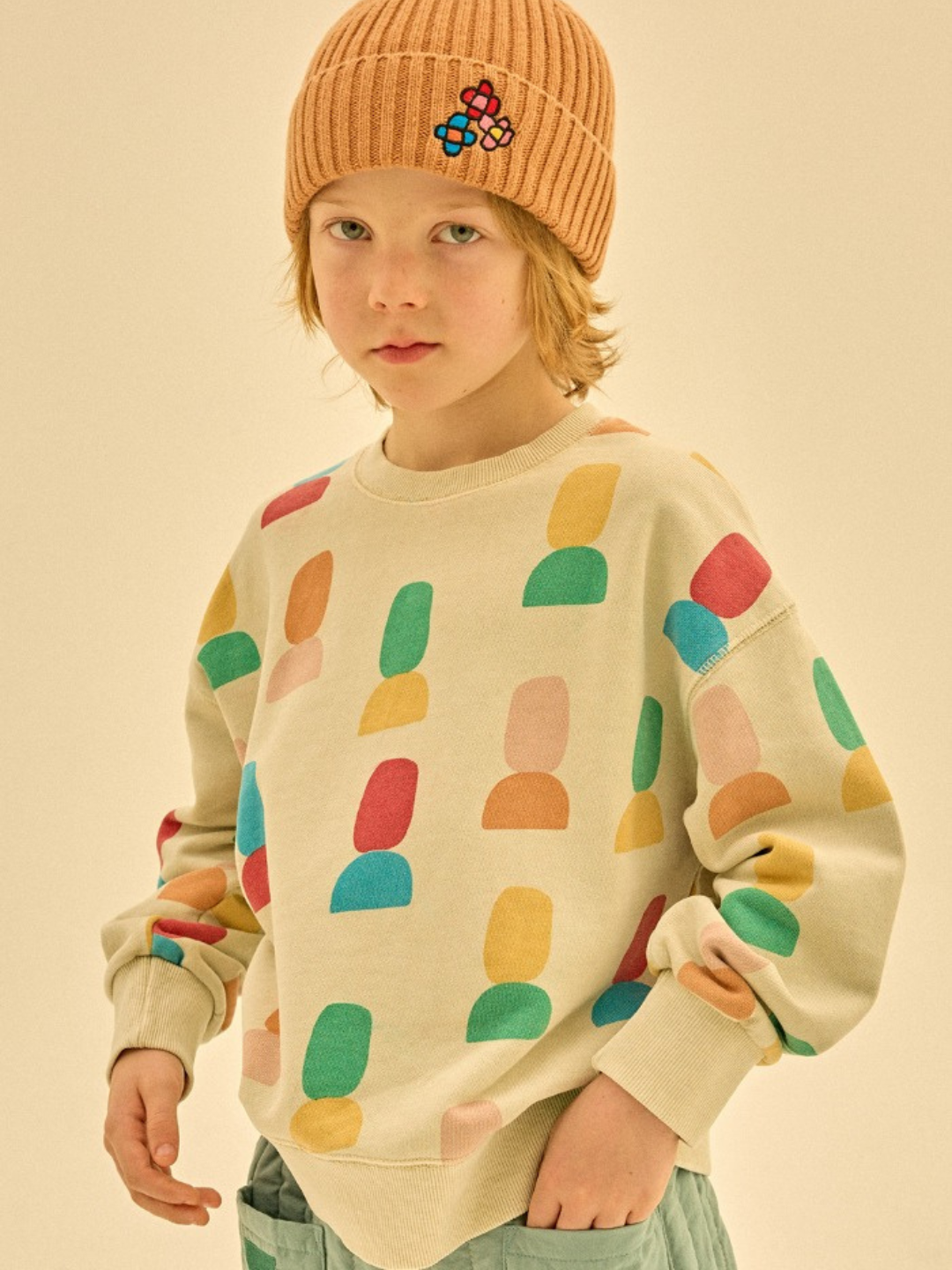 A child wears a vibrant, patterned sweatshirt featuring the SHAPE PIGMENT SWEATSHIRT, complemented by a soft rust-colored knit hat. The plain beige background accentuates the meticulous merchandising.