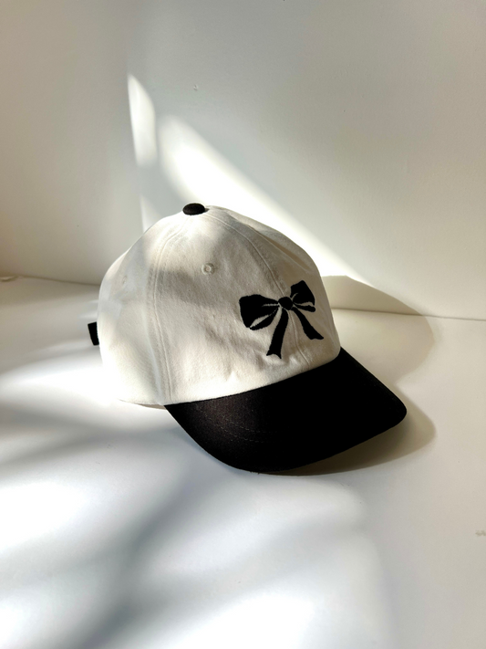 Image of A front view of the kid's Bow Cap. White hat with black cap and black bow embroidery in the center. 
