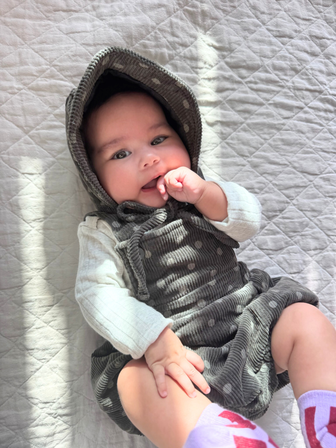 A baby wearing the DOT ROMPER + BONNET SET, a charcoal polka dot cotton corduroy overall onesie and a matching bonnet. She wears purple and burgundy striped socks, and is lying on a beige quilt.