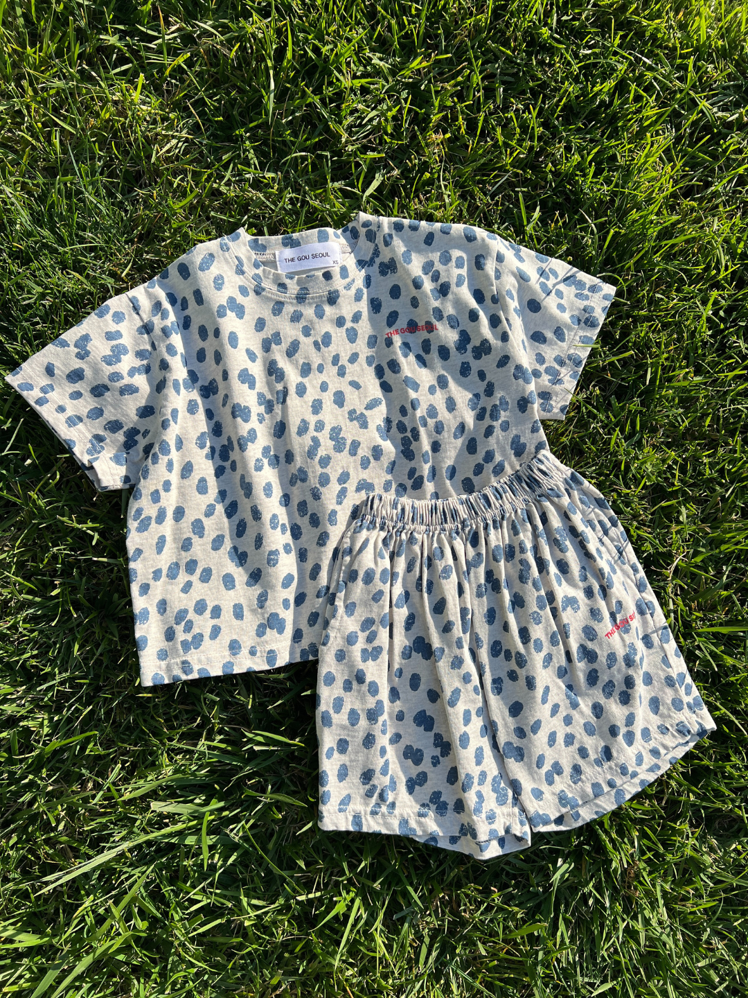 A matching children’s set of the SPOTTED TEE short-sleeved cotton jersey shirt and shorts, featuring a grey base with a blue polka dot pattern, embroidered with the phrase "je suis cool," neatly laid out on green grass. This Korean kids brand guarantees comfort and style for your little one.