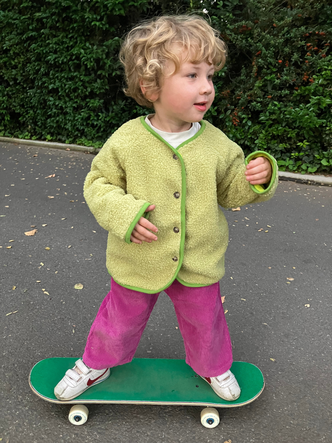 Matcha | A toddler wearing a light green collarless fleece jacket with four brown buttons. He stands on a green skateboard, and wears magenta pants and white sneakers, on a grey asphalt path with a green hedge behind.