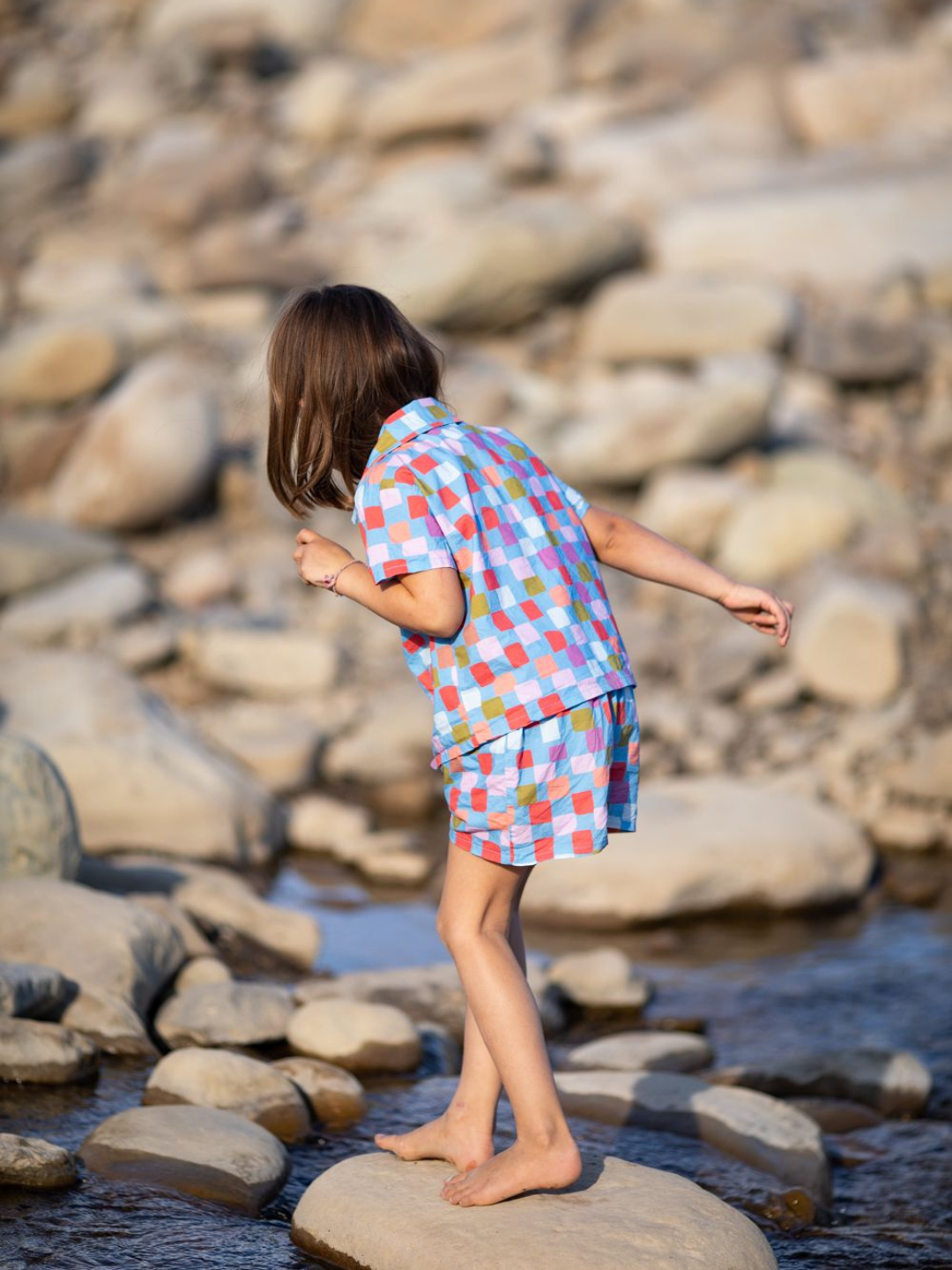 Blue | A child walking over stepping stones wearing a kids' shirt and shorts set in a pattern of red, orange, pink, lilac and green squares on a blue background